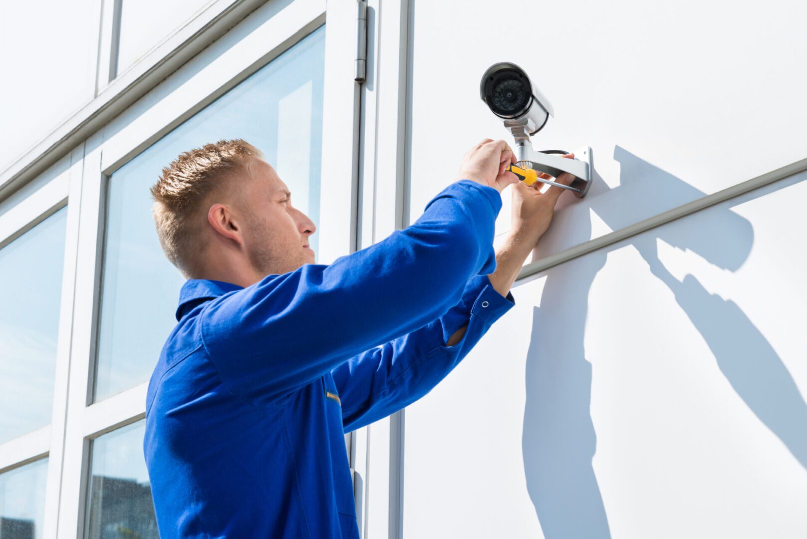 A person in a blue uniform uses a screwdriver to install a security camera on the exterior wall of a building.