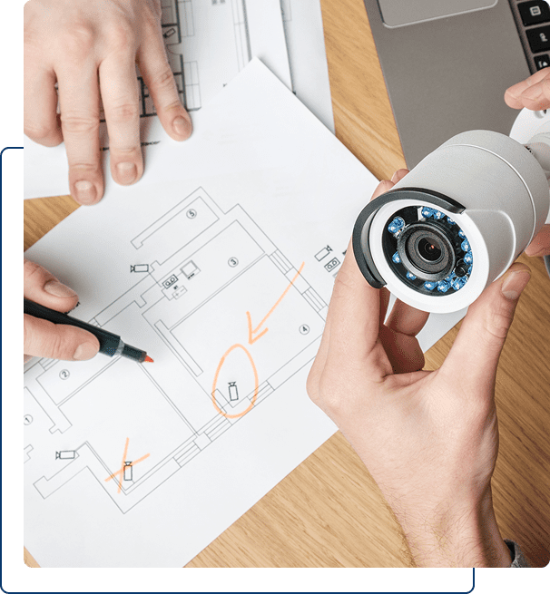 Two people review a building floor plan with marked locations while holding a security camera, discussing surveillance placement.